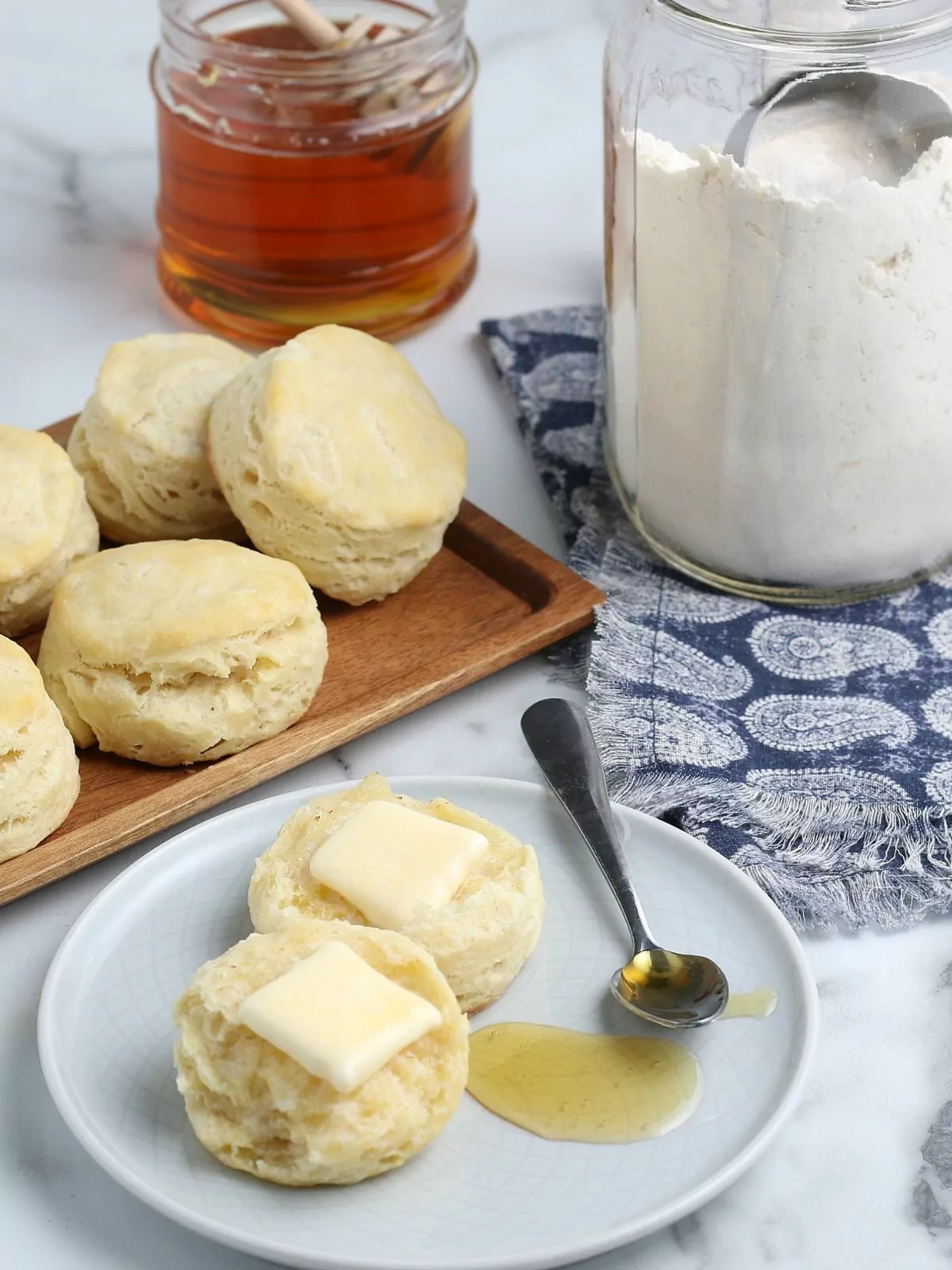 biscuit split in two with pats of butter on each half and spoon of honey on the plate. Jar of homemade bisquick and a jar of honey with the rest of the biscuits are in the background.