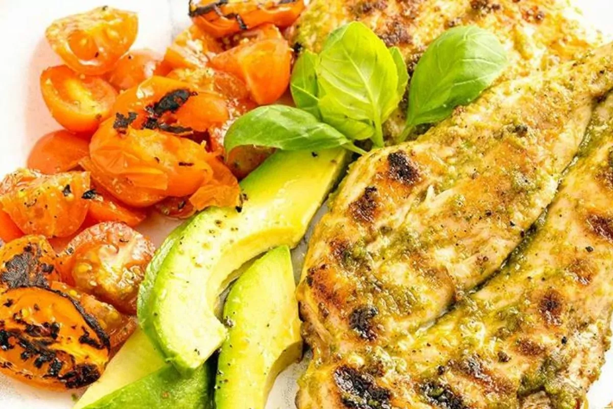 A plate of pesto coated grilled chicken with avocado and grilled tomatoes.