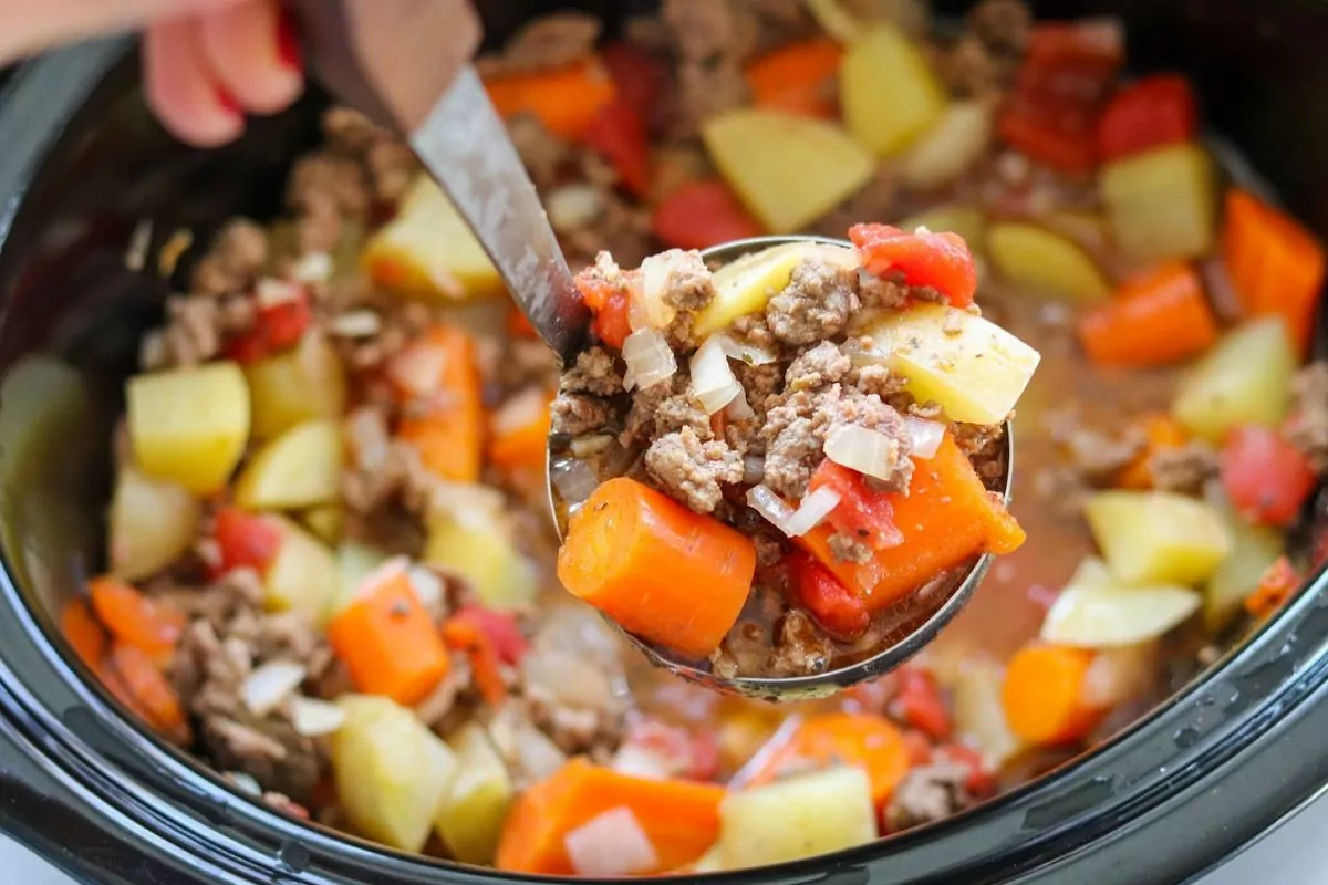 a pot of stew with a ladle lifting burger, potatoes and carrots.