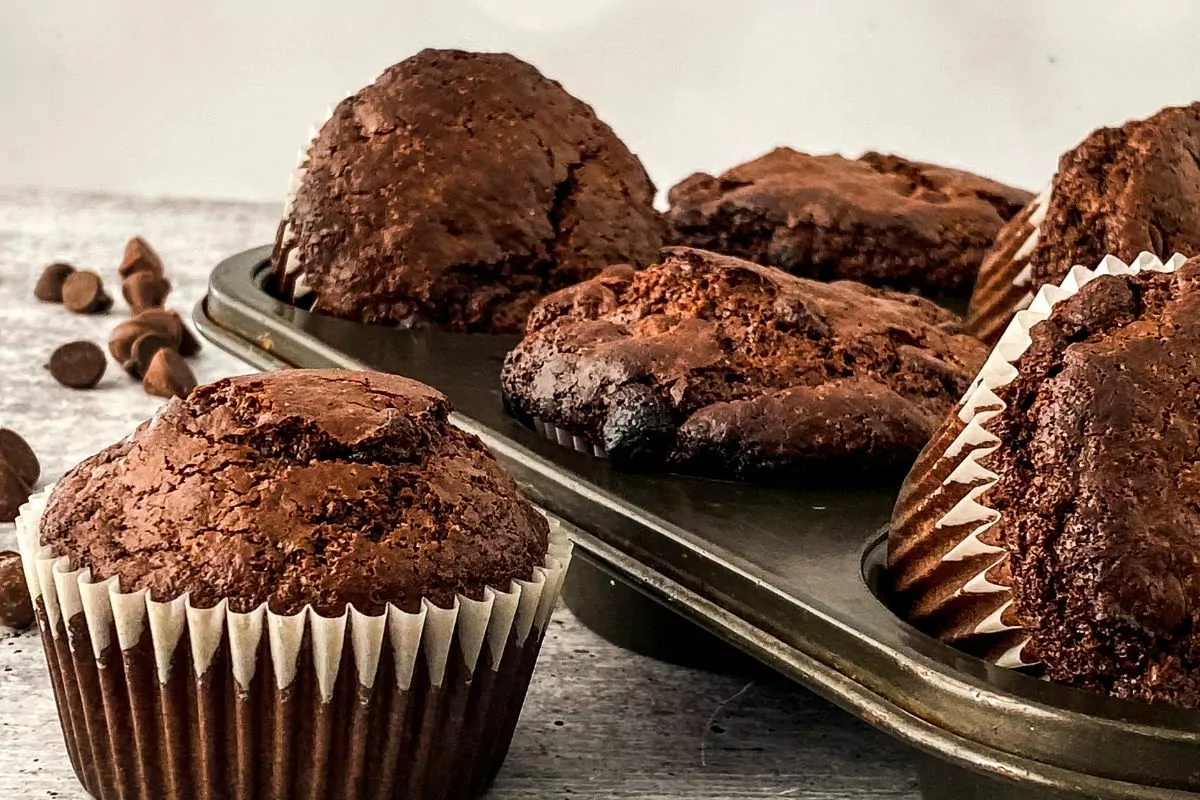 chocolate muffins with chocolate chips in muffin pan and one in wrapper on counter.