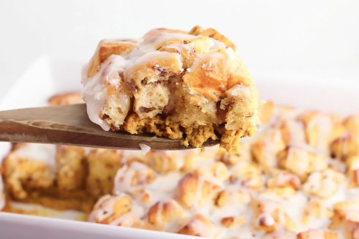 slice of Pumpkin Cinnamon Roll Casserole on a wooden spatula taken out of the casserole in the background