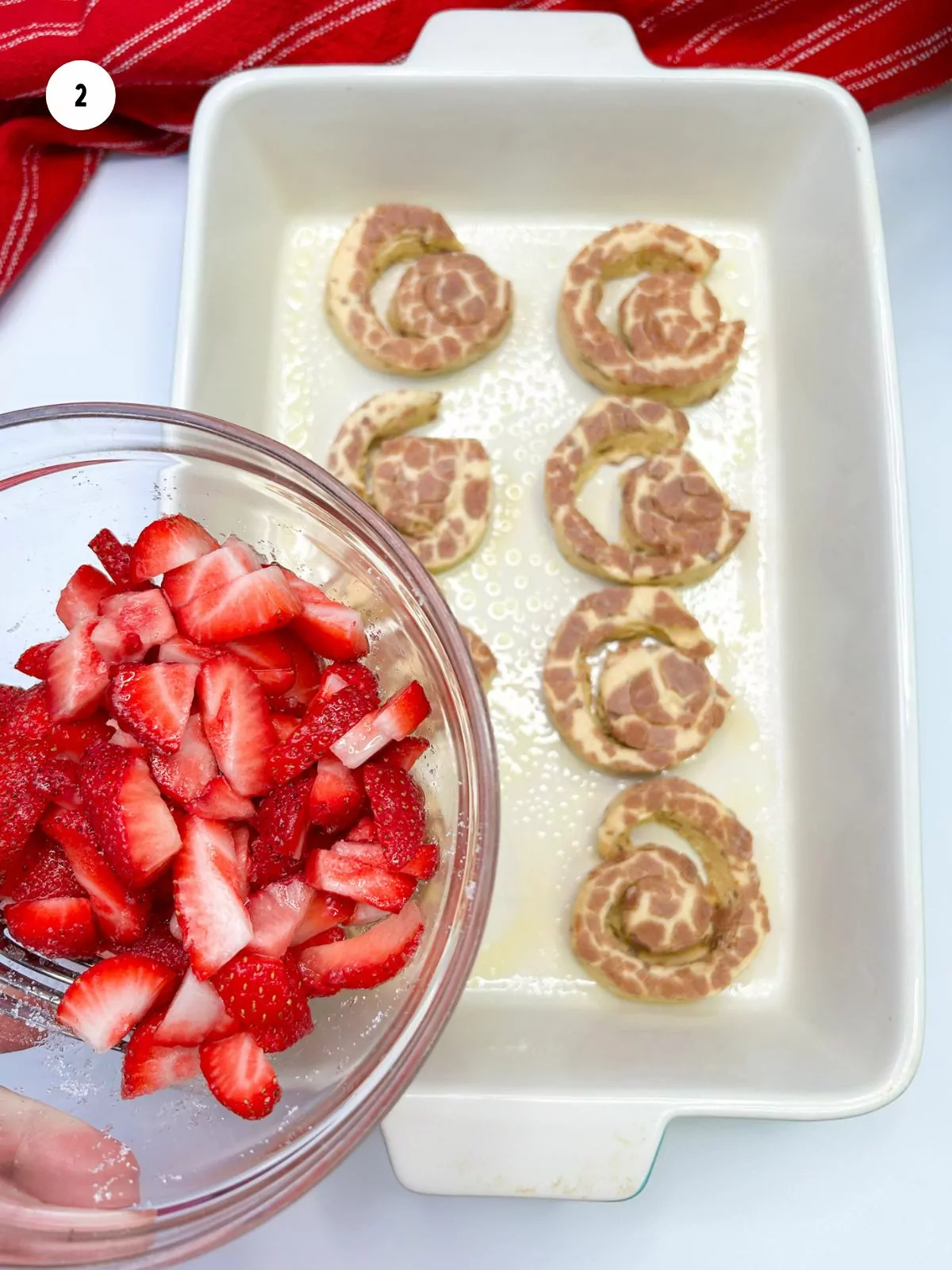 sliced strawberries in bowl being held over casserole dish with cinnamon rolls.