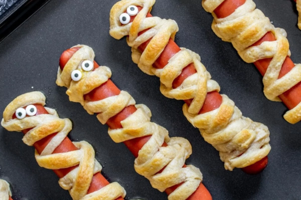 a line of hot dogs, wrapped in dough, and adorned with eyes to make the perfect halloween dinner.