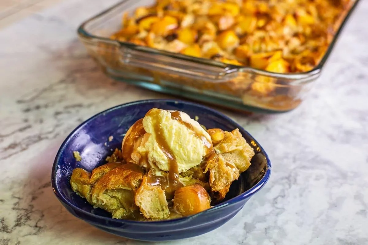 A plate of bread pudding made with peaches. A scoop of ice cream rests on top with caramel sauce drizzle.