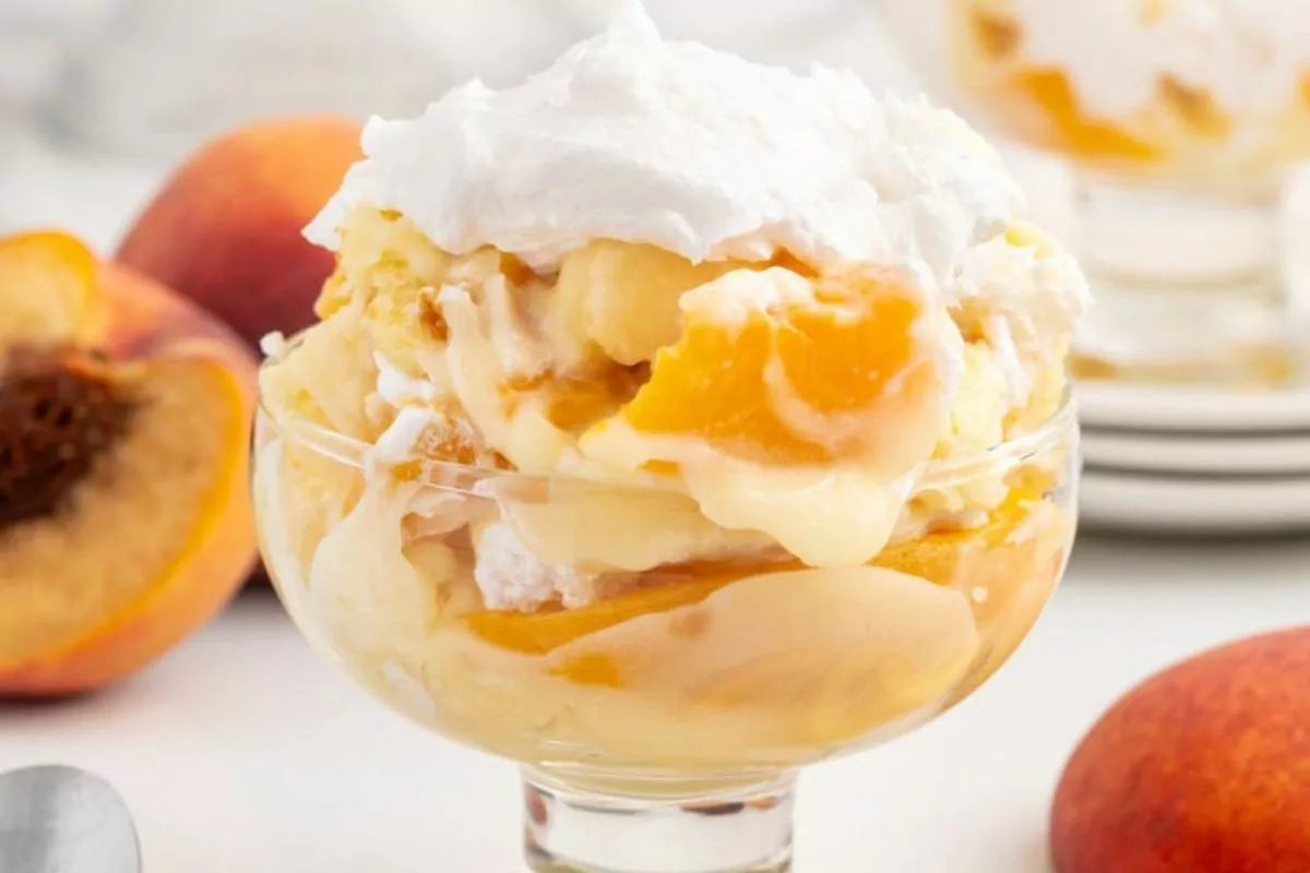 A glass bowl filled with peaches and cream pound cake trifle, with pudding and whipped cream.