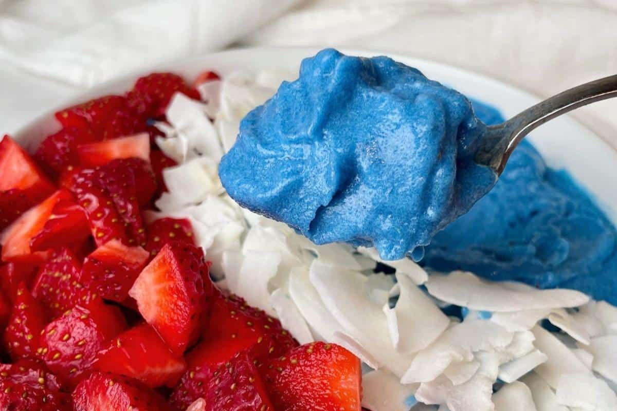 A blue peach smoothie bowl with berries and coconut, with a bite lifting away with a spoon.