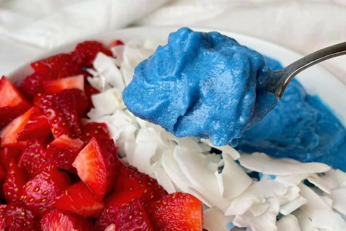 A blue peach smoothie bowl with berries and coconut, with a bite lifting away with a spoon.