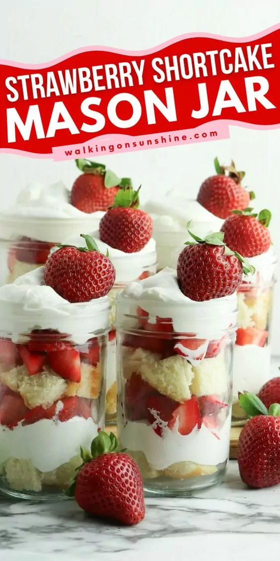 mason jar with cake, strawberries and whipped cream layered with whole strawberries scattered around.