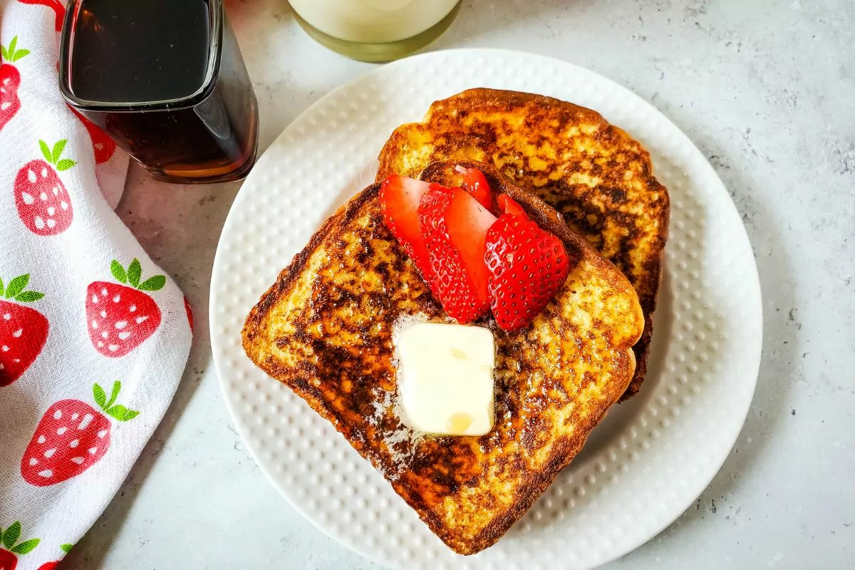 two slices of French Toast on white plate with a pat of butter, syrup and strawberries.