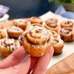 hand holding a mini cinnamon roll over a tray of more.