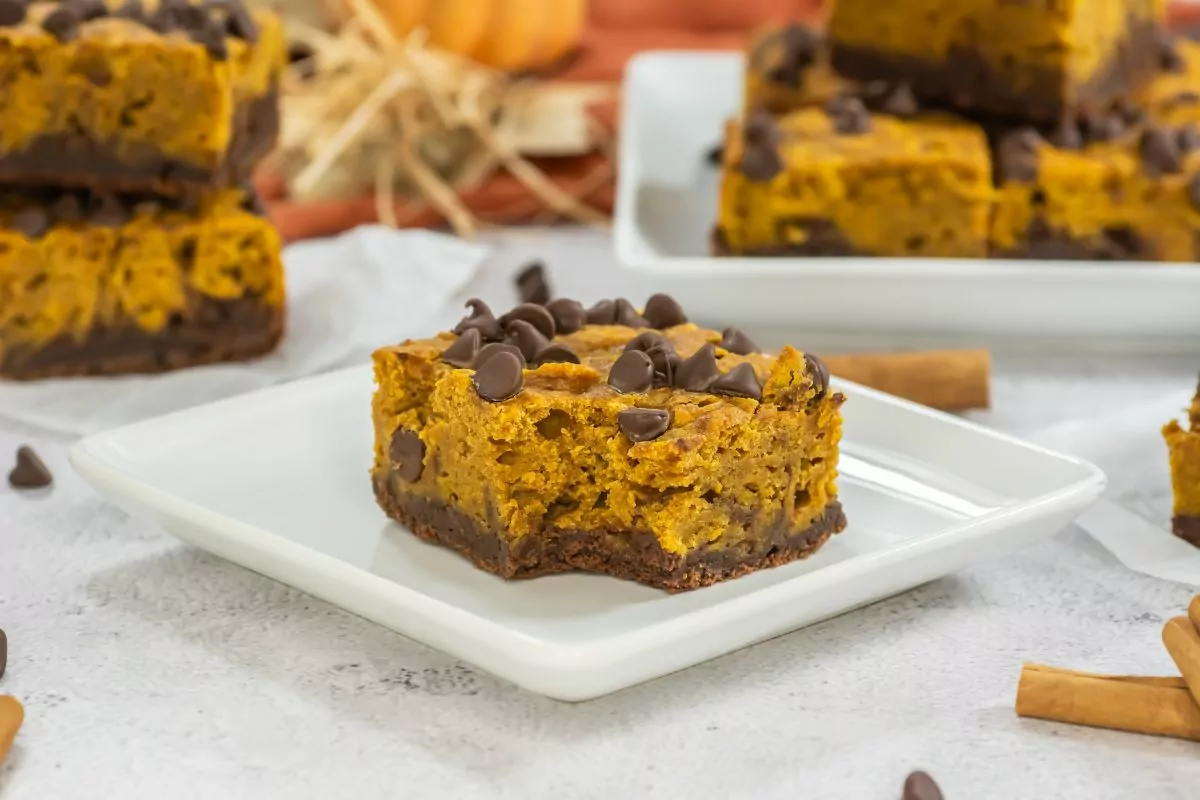 pumpkin brownie with a bite taken out served on a square white plate with the rest on a platter in the background.