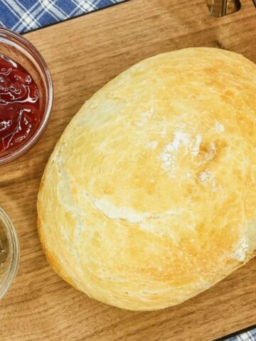 loaf of oval shaped baked bread on cutting board with bowl of jam and butter.