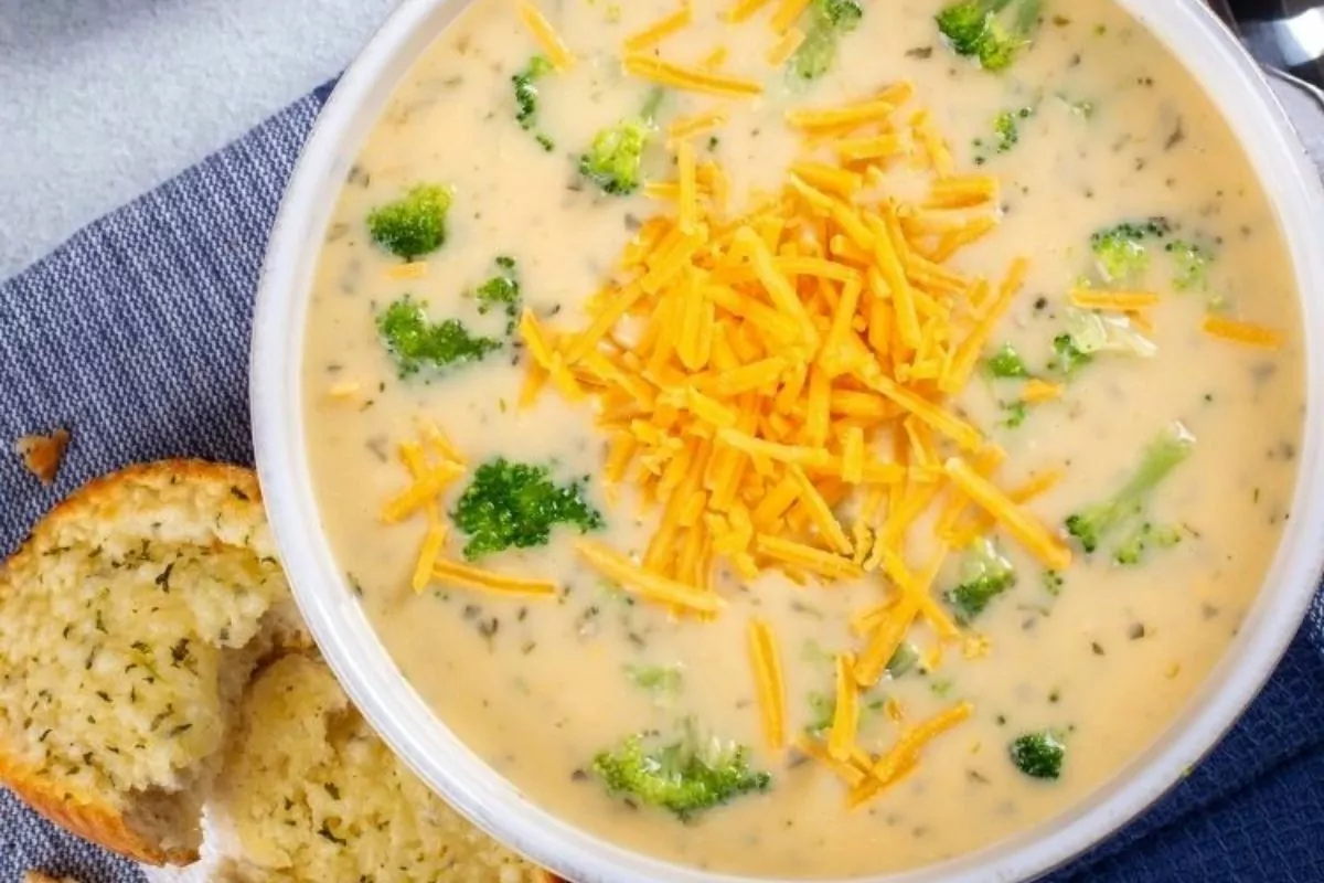 a bowl of creamy soup with cheddar cheese and broccoli. A piece of half-eaten bread is nearby.