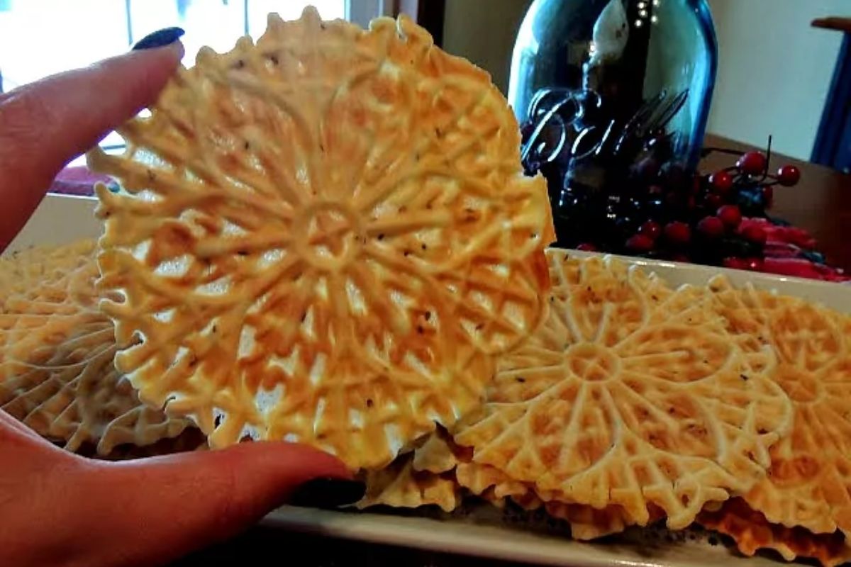 a hand holding a fresh baked pizzelle cookie over a tray of cookies.