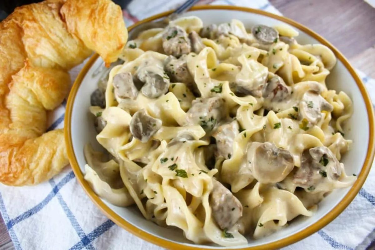 a bowl of beef chunks, noodles and creamy sauce with croissant bread