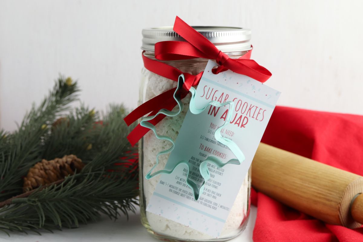 sugar cookie mix in a jar with cookie cutter and gift tag attached. Sprig of pine and rolling pin in foreground.