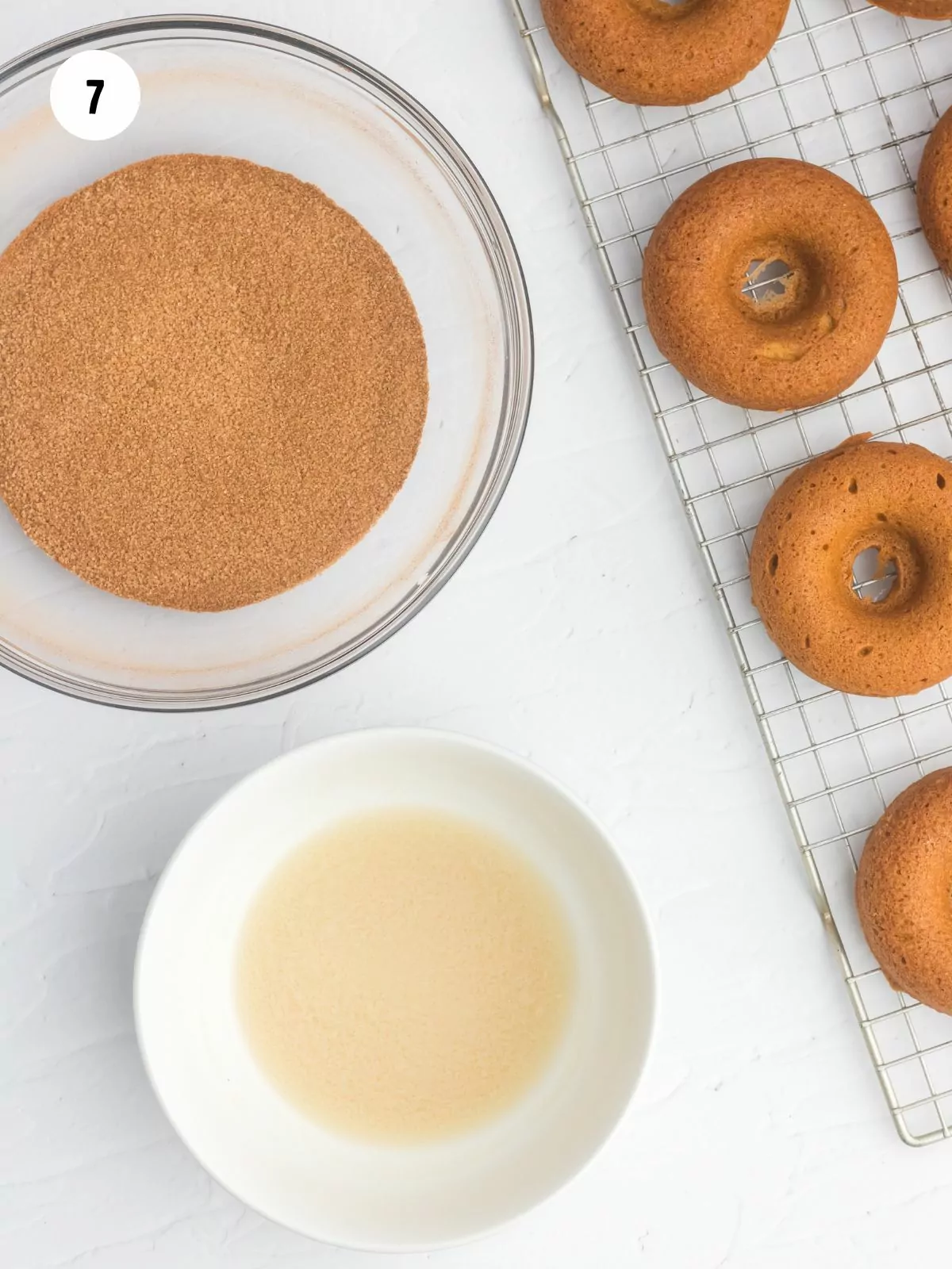 top bowl filled with cinnamon sugar. Bottom bowl filled with melted butter. To the right is a cooling rack with pumpkin donuts on it