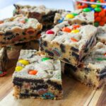 Cookie bars with colorful M&M's, Oreos, and peanut butter cups piled on a wood board