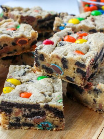 Cookie bars with colorful M&M's, Oreos, and peanut butter cups piled on a wood board