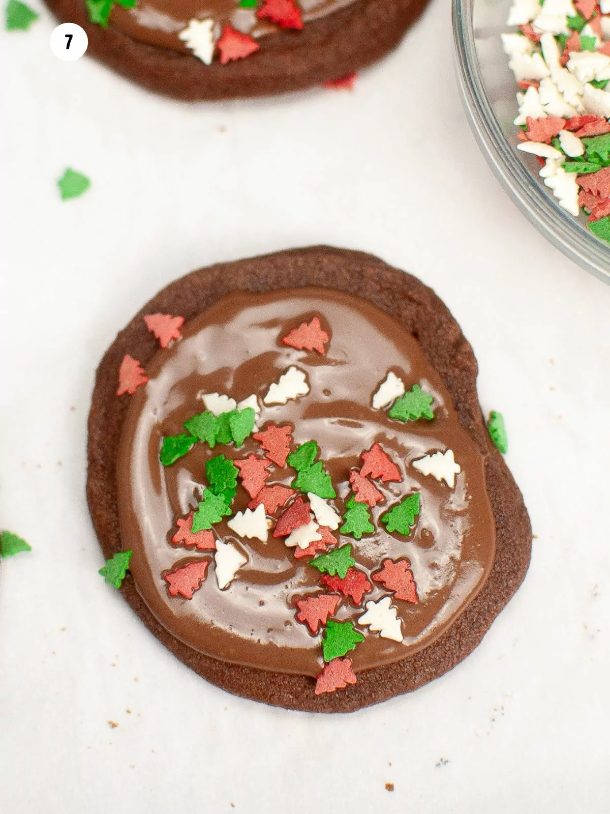 overhead view of chocolate cookies with melted chocolate and Christmas tree sprinkles on parchment paper.