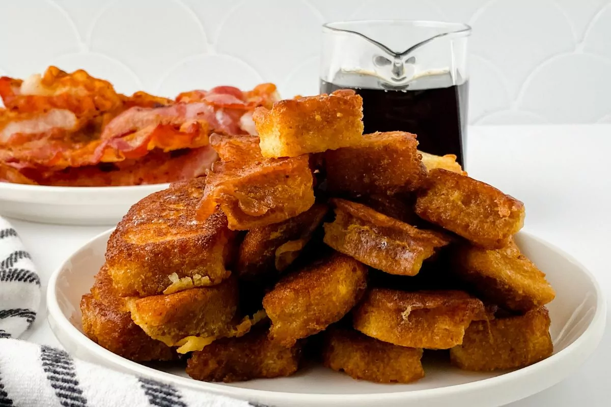 plate of French Toast Sticks with a plate of bacon in the background.