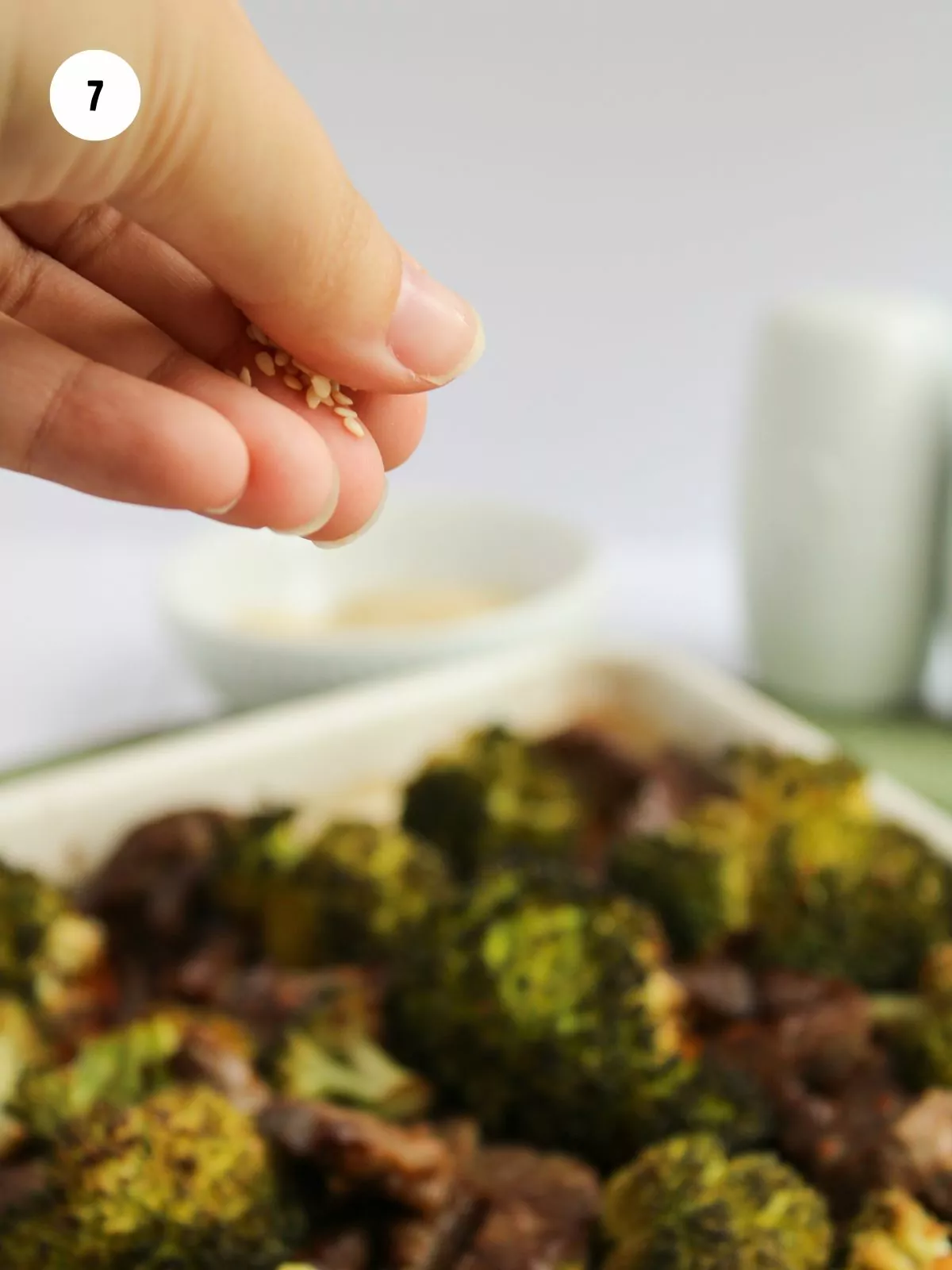 hand holding sesame seeds to sprinkle over baked beef and broccoli