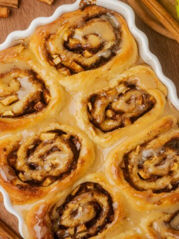 Apple Filled Cinnamon Rolls in a white scalloped edge baking dish. Green apples and cinnamon sticks on the table next to the baking dish