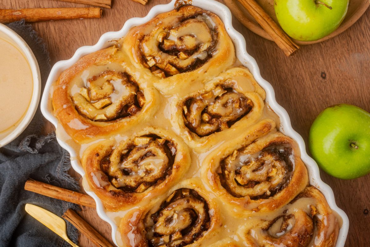 Apple Filled Cinnamon Rolls in a white scalloped edge baking dish. Green apples and cinnamon sticks on the table next to the baking dish