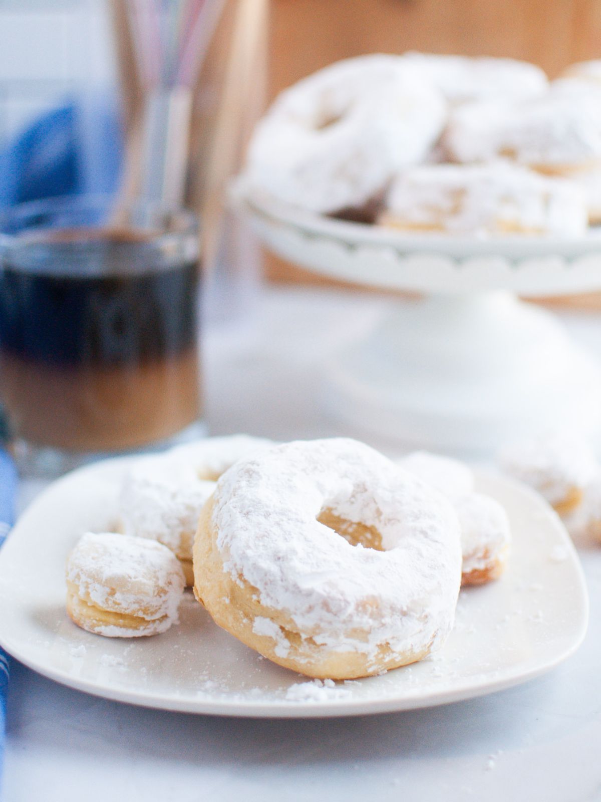 Air Fryer Biscuit Donuts with Powdered Sugar on a plate and a cake stand full in the background