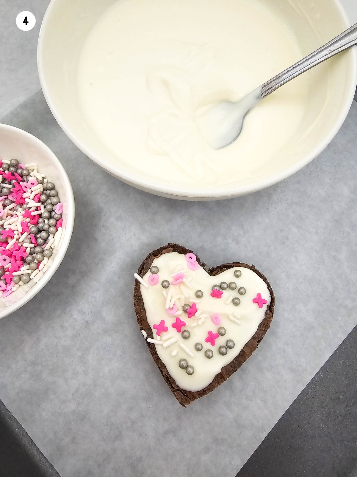 melted white chocolate in bowl with heart shaped brownie decorated on parchment paper.