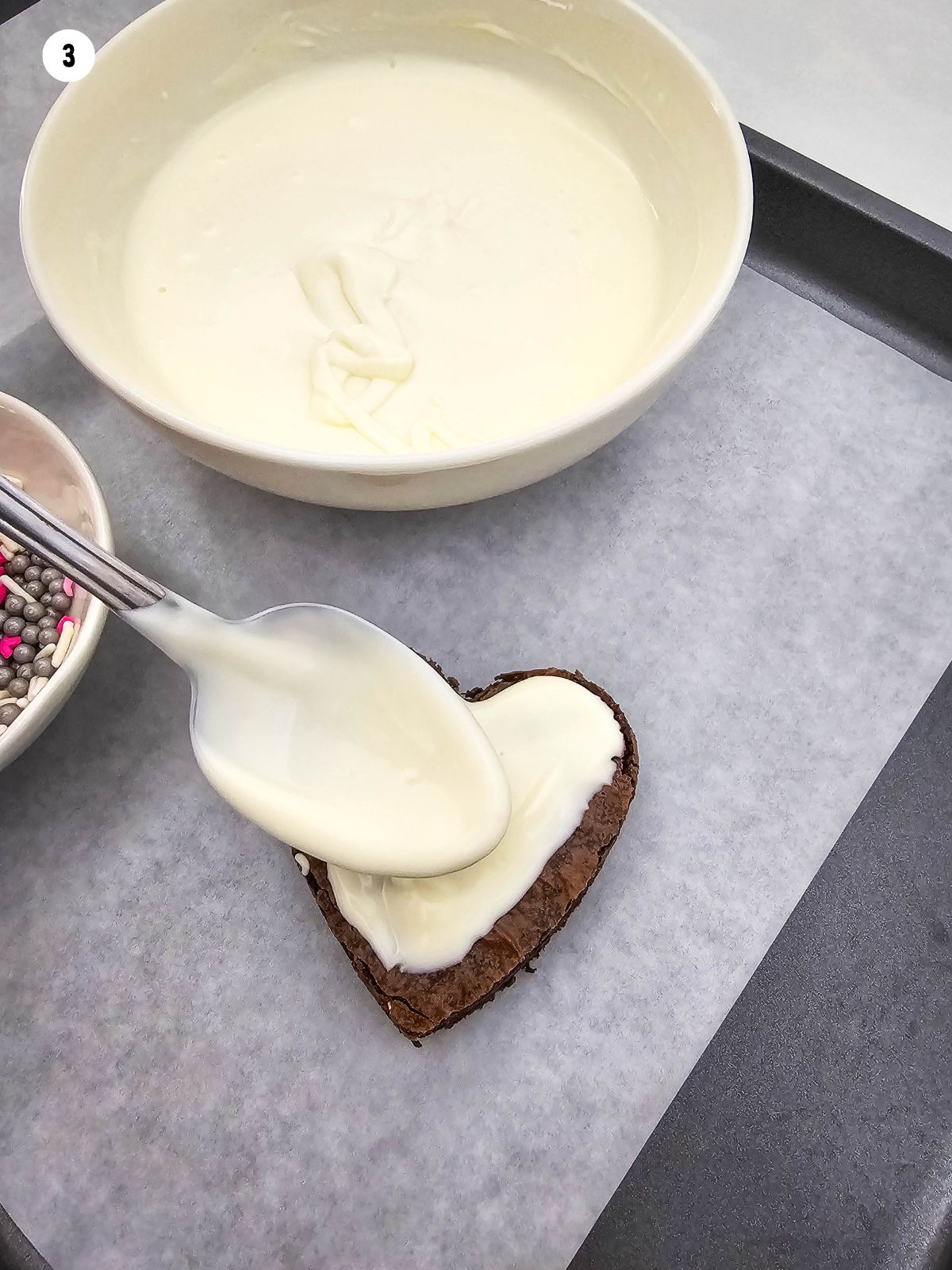 melted white chocolate in bowl and being spread on top of brownie with spoon.