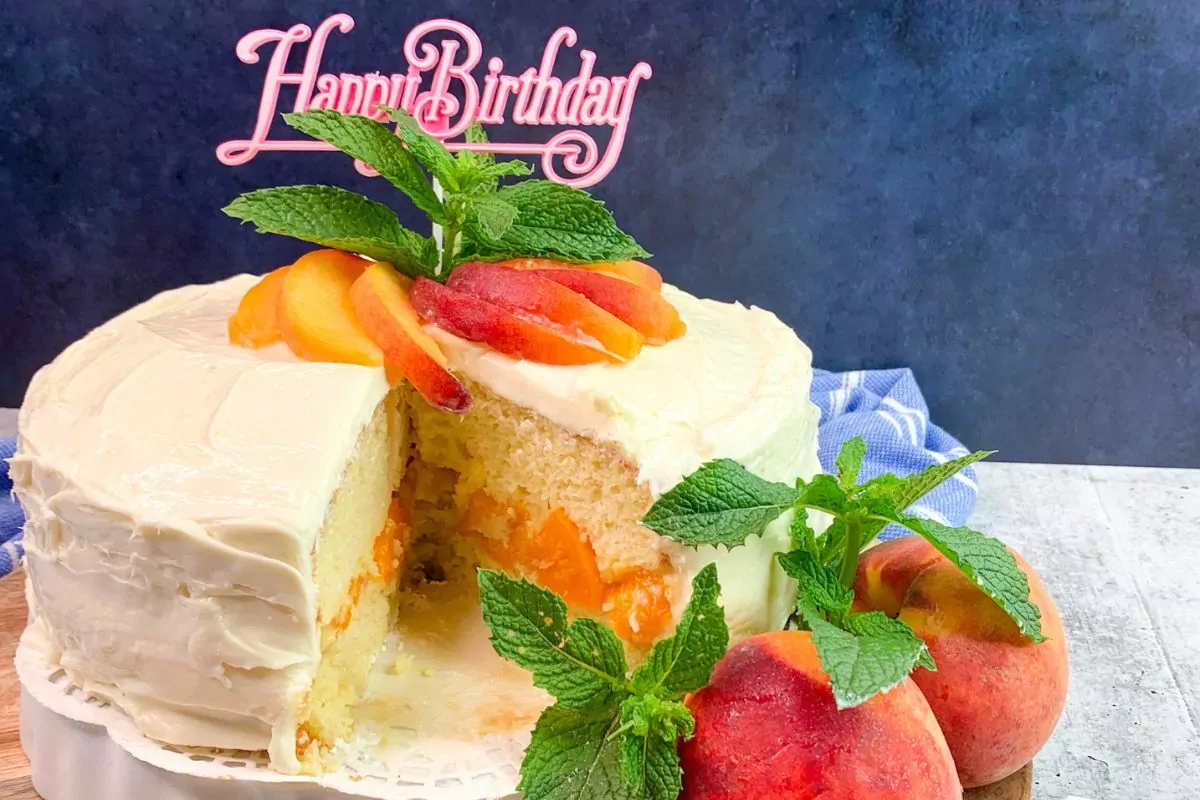 peach birthday cake sliced and on cake server with happy birthday sign, fresh peaches and fresh mint.