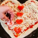 Pepperoni cut out into heart shapes being placed on uncooked pizza.