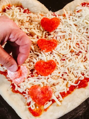Pepperoni cut out into heart shapes being placed on uncooked pizza.