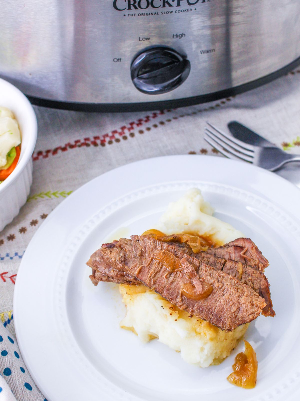 slices of flank steak and onions served over mashed potatoes on a white plate. The slow cooker is in the background