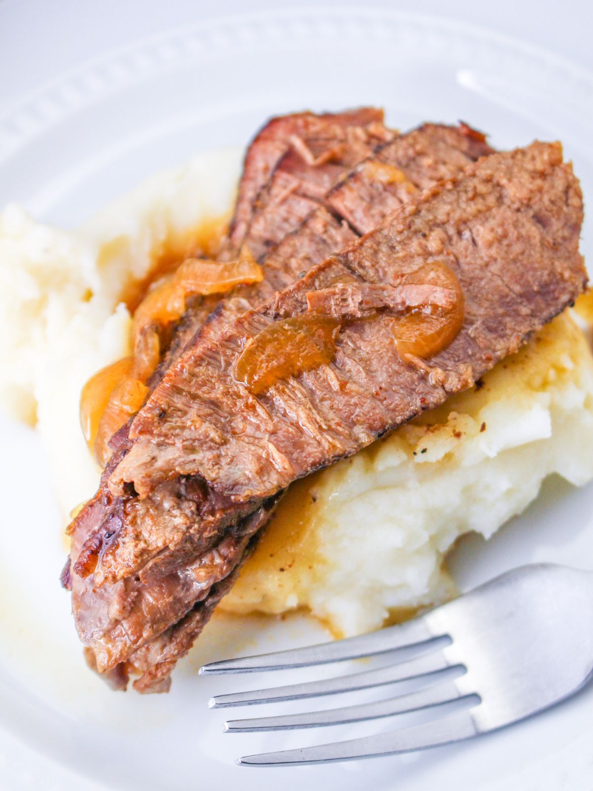 slices of flank steak and onions served over mashed potatoes on a white plate with a fork