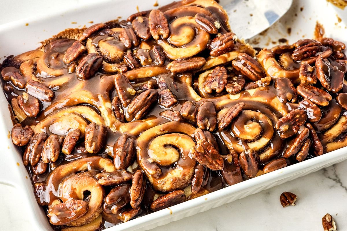 Butter Pecan Cinnamon Rolls in a white casserole dish with a pie server lifting a roll out.