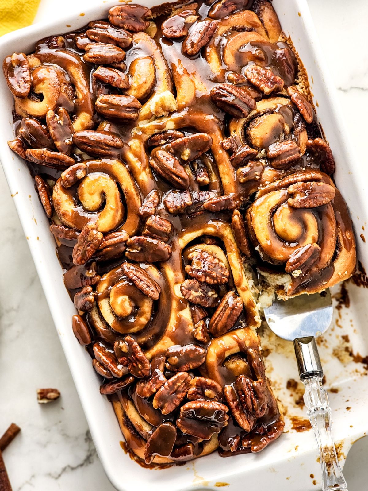 Butter Pecan Cinnamon Rolls with a pie server lifting a roll out of the dish