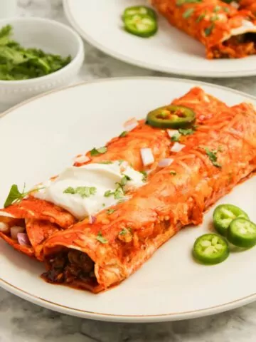 two Cheesy Beef Enchiladas topped with diced red onion, sour cream, and slice jalapeno served on a white plate.