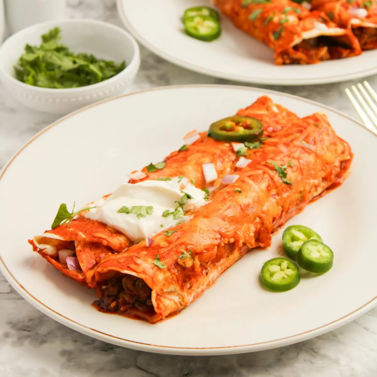 two Cheesy Beef Enchiladas topped with diced red onion, sour cream, and slice jalapeno served on a white plate.