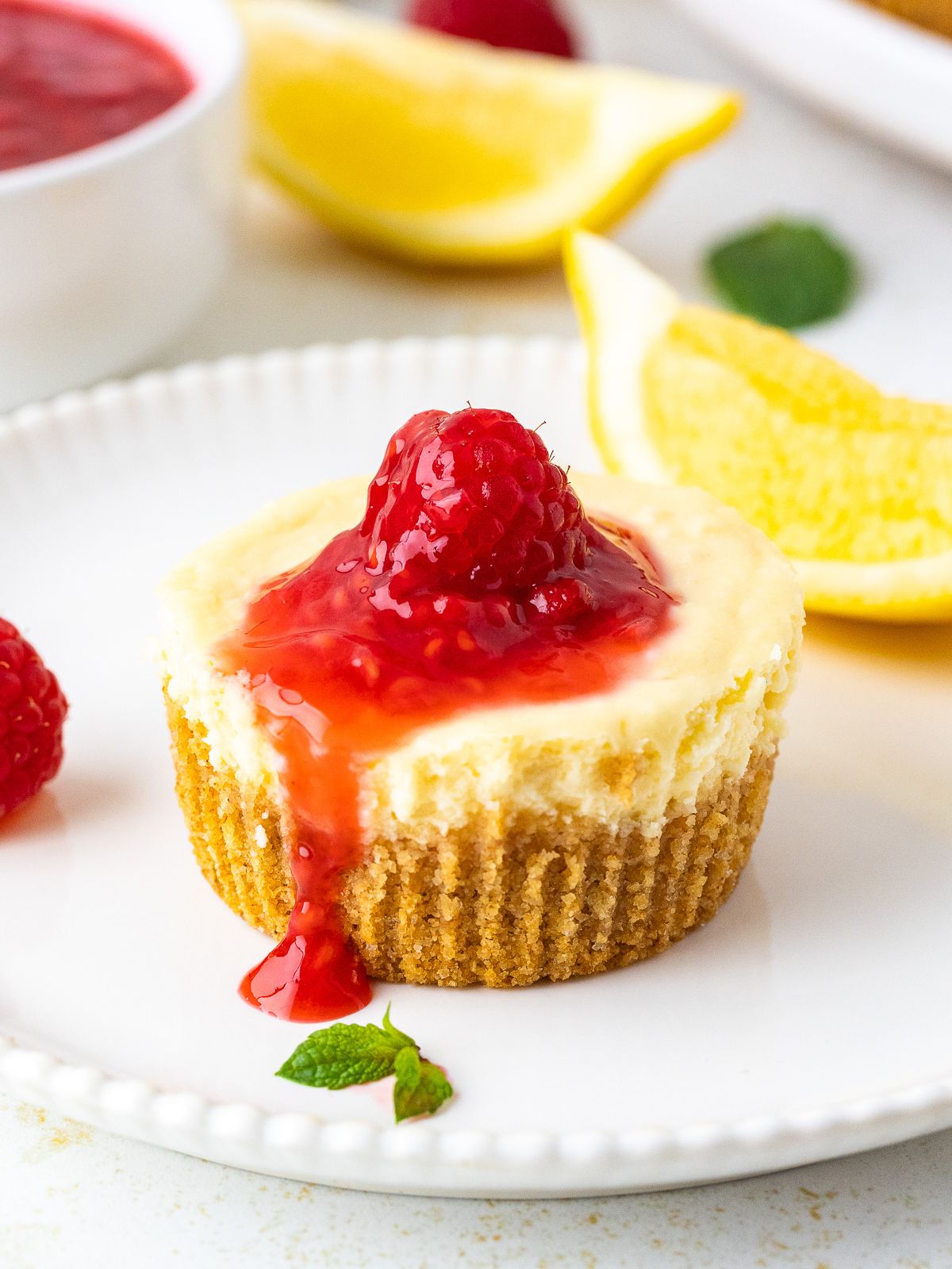 Raspberry Mini Cheesecakes with raspberry sauce dripping down the side and lemon slices in the background