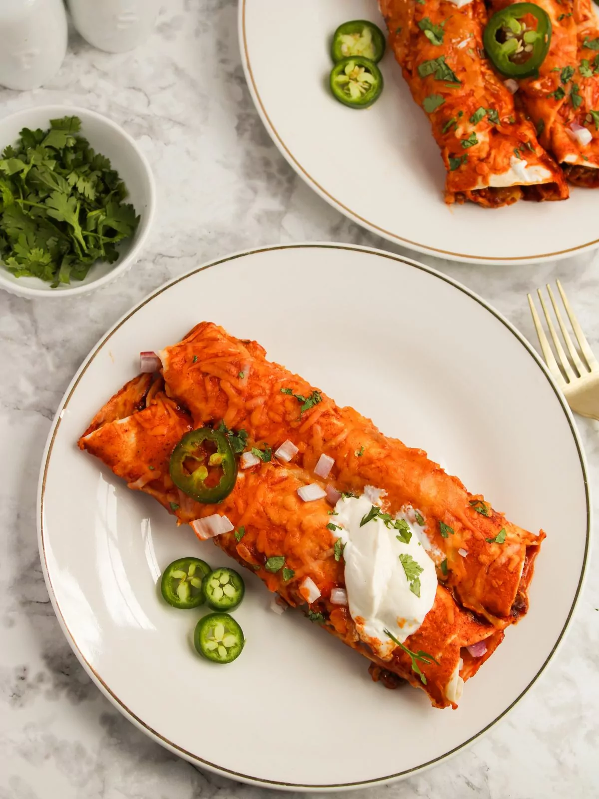 overhead shot of Cheesy Beef Enchiladas served on a white plate with fresh cilantro, sour cream, onion, and jalapeños.