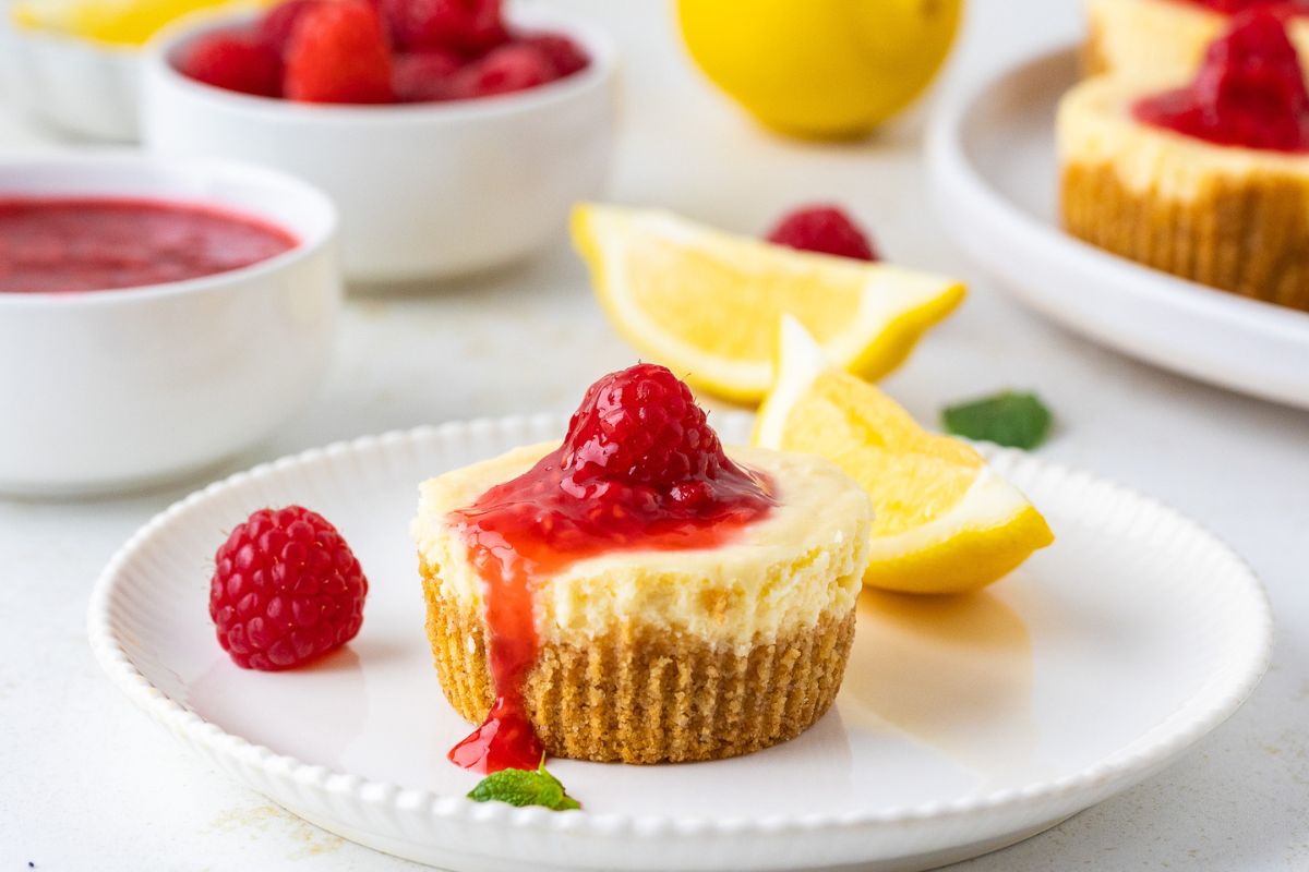 Mini Cheesecakes topped with raspberry sauce dripping down the side. Lemon slices and a bowl of fresh raspberries in the background