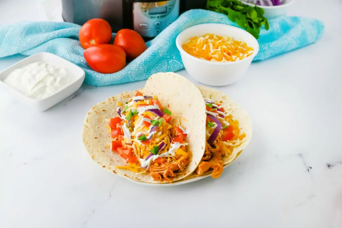 Two shredded chicken tacos on a plate with toppings in the background