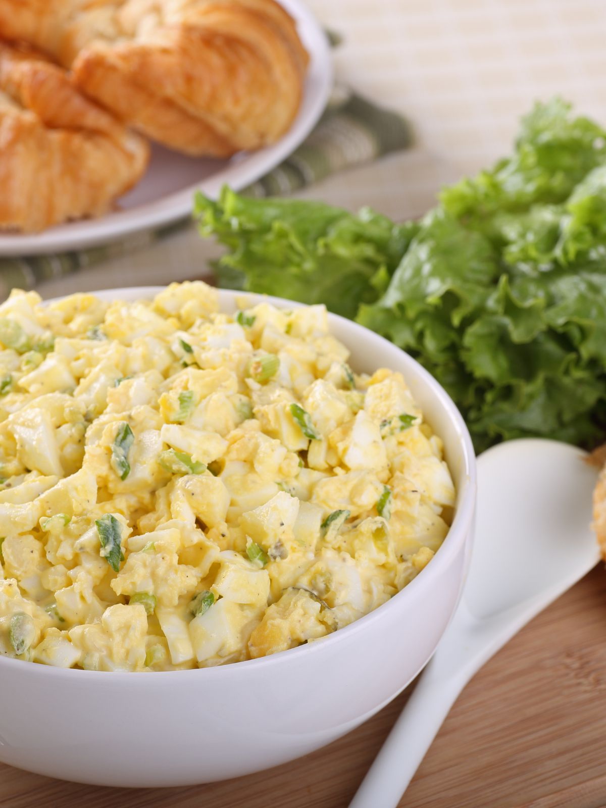 egg salad in white bowl with lettuce and croissants in the background.