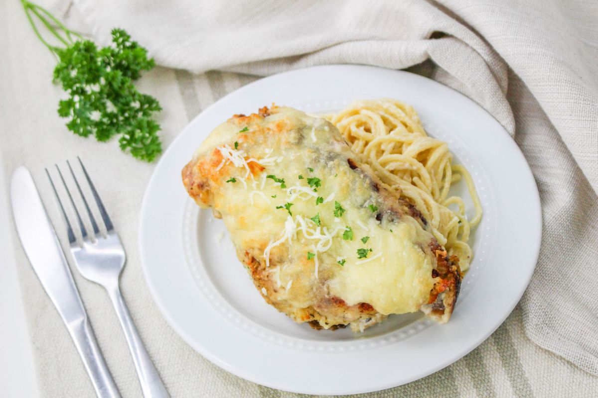 Baked Chicken Cutlet Alfredo served with pasta on a white plate with cutlery next to the plate