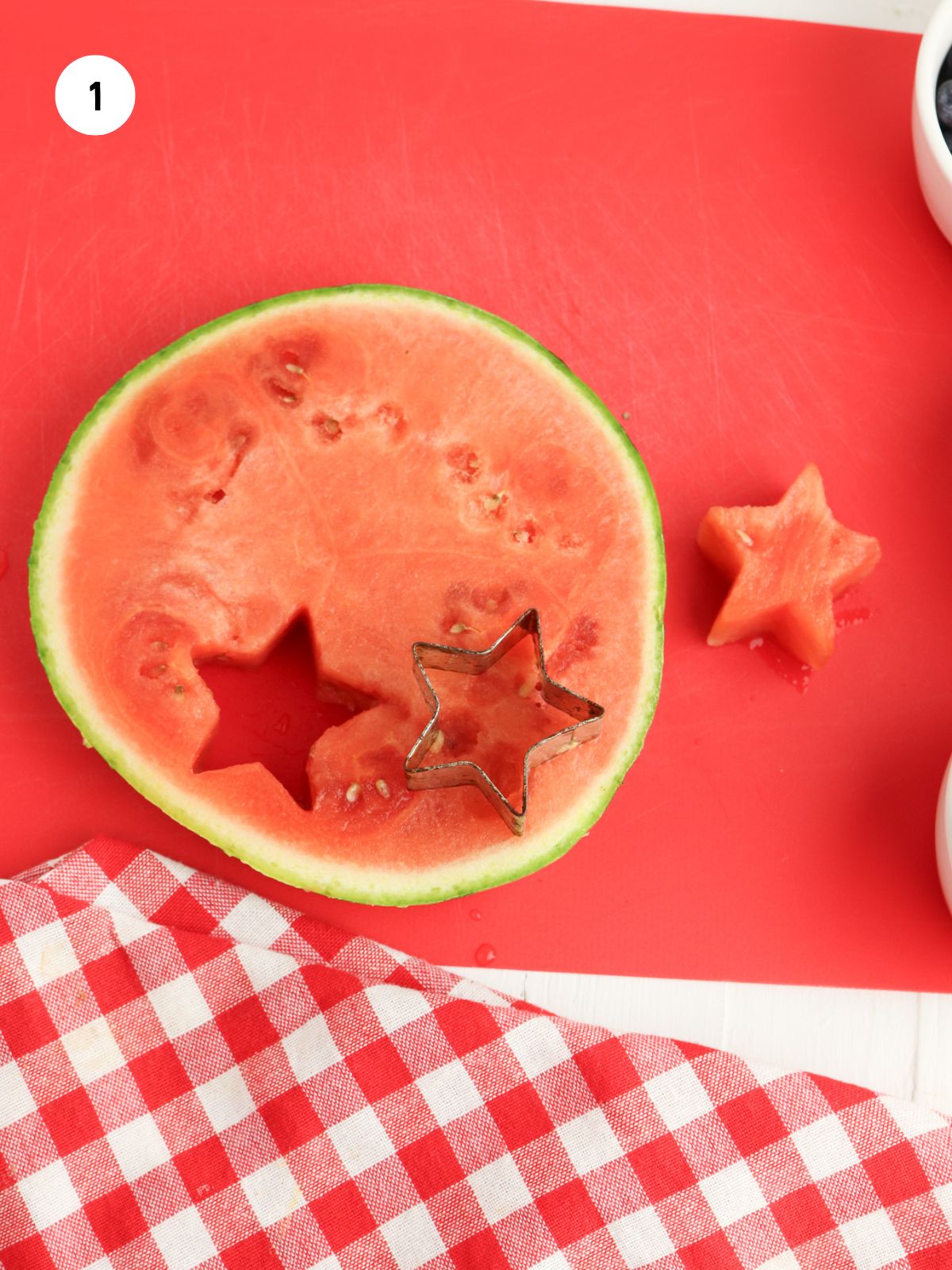 Round slice of watermelon on a red cutting board. A mini star cookie cutter is cutting star shapes out of the watermelon.