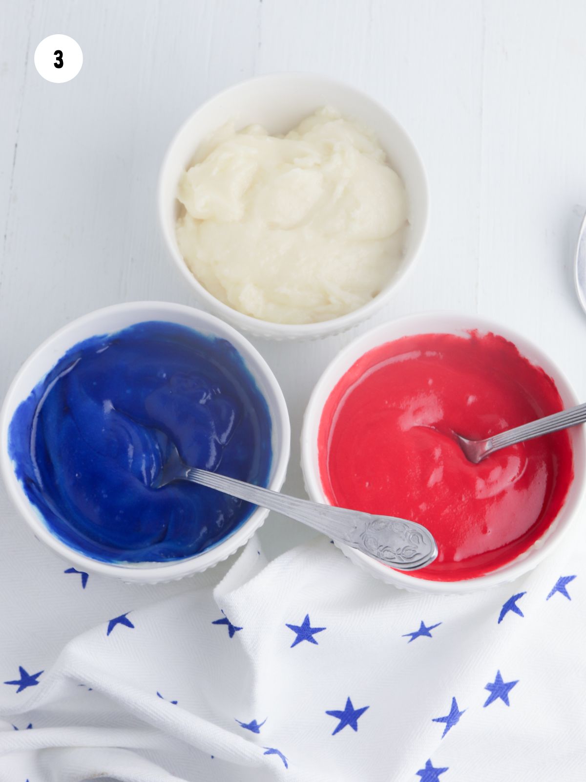 Three small bowls filled with white, blue, and red frosting.
