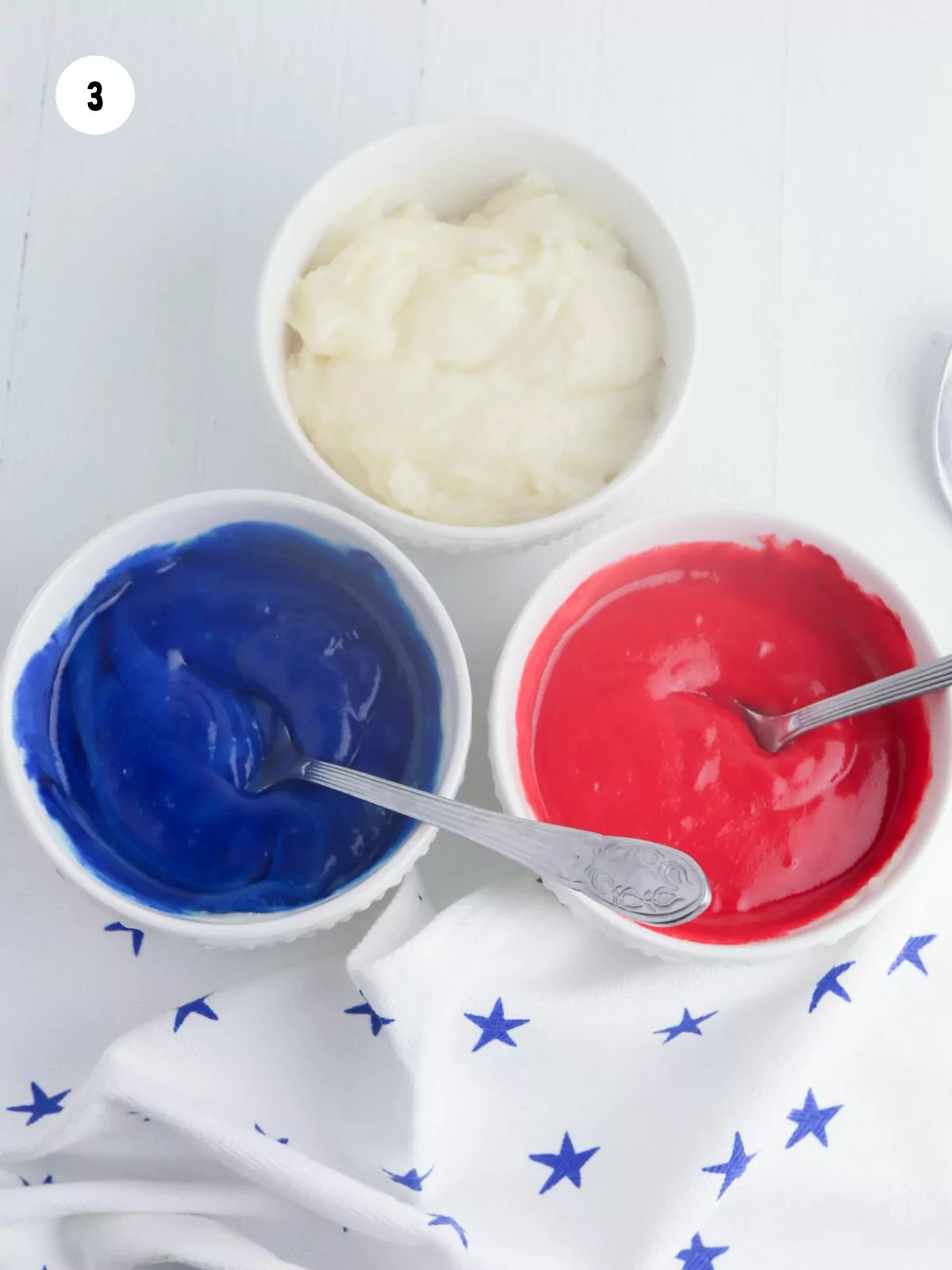 Three small bowls filled with white, blue, and red frosting.