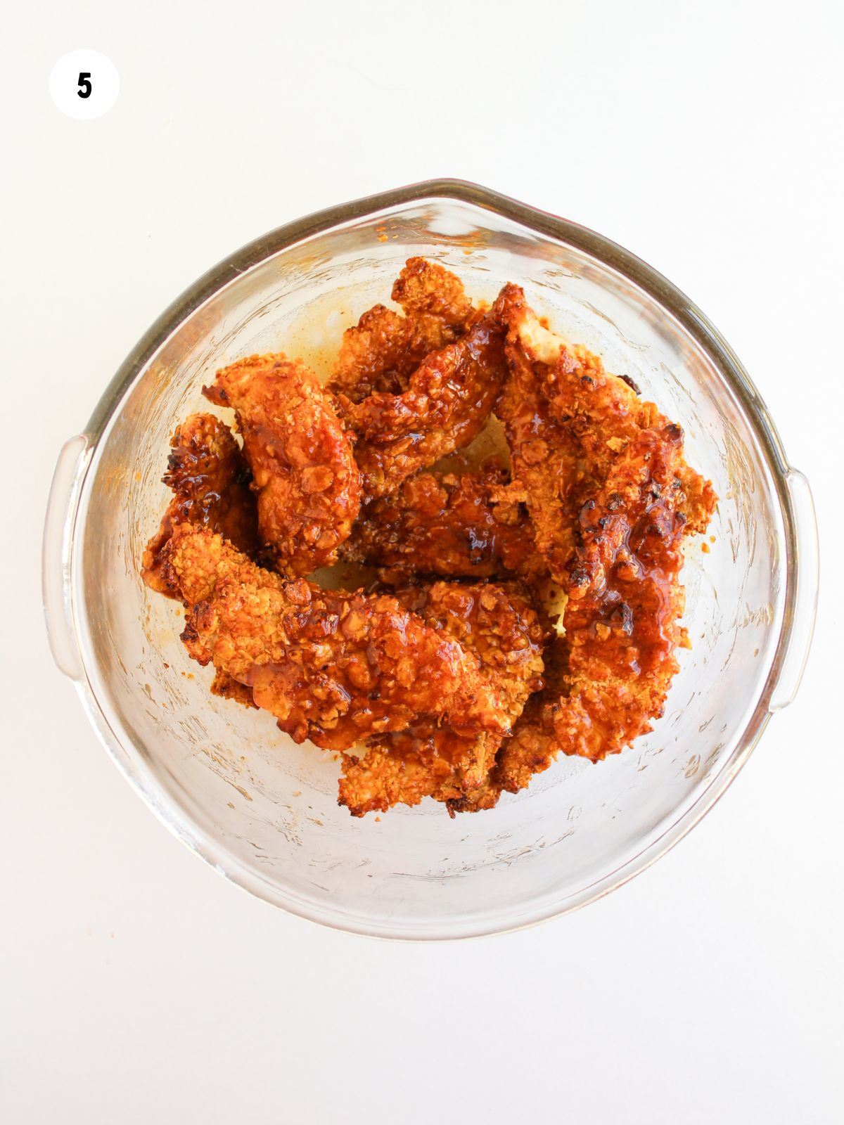 Chicken tenders tossed in honey bbq sauce in a clear mixing bowl.
